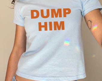 Könnte beinhalten: Hellblaues T-Shirt mit dem orangefarbenen Text "DUMP HIM". Das Shirt hat einen Rundhalsausschnitt. Das Model trägt auch eine blaue karierte Shorts. Das T-Shirt scheint aus einem weichen Material zu sein.