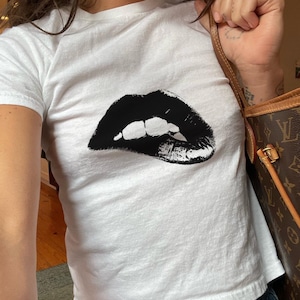 Pode incluir: Camiseta branca com um gráfico preto de uma boca grande aberta com dentes.
