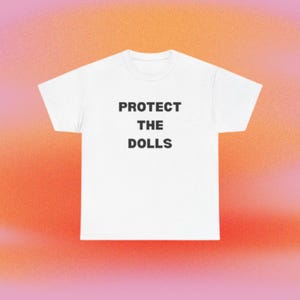 Może przedstawiać: Biała koszulka z czarnym napisem "PROTECT THE DOLLS". Koszulka jest umieszczona na gradientowym tle w odcieniach pomarańczu i różu.