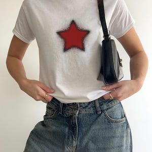 May include: White t-shirt with a red star design and black edging. The shirt is worn with blue denim jeans and a black shoulder bag. The star graphic is a prominent feature on the front of the tee.