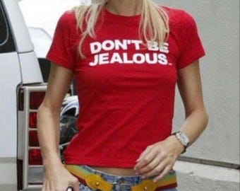 May include: A woman wearing a red t-shirt with the text "DON'T BE JEALOUS."  She is also wearing a yellow belt, blue denim skirt, and sunglasses. She is holding a pink handbag with a large number 7 on it.