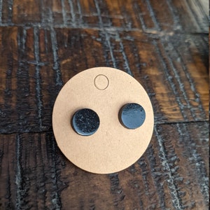 May include: A pair of black stud earrings displayed on a brown card. The earrings are round and have a slightly textured surface.