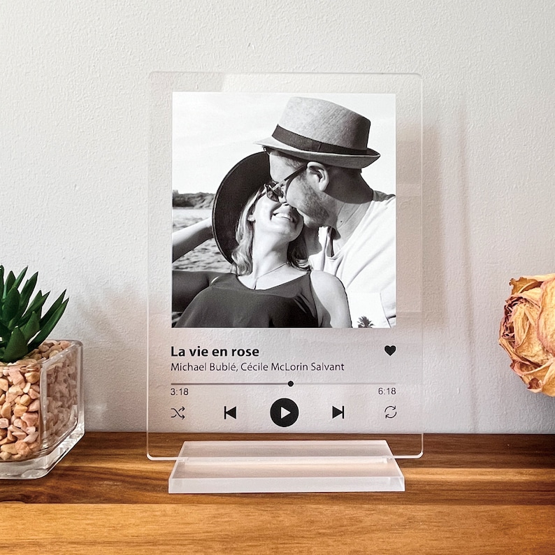 May include: Clear acrylic photo display featuring a black and white image of a couple kissing. The display includes the song title "La vie en rose" and artist information. A clear stand supports the display, with a small plant and dried flower in the background.