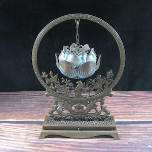 Könnte beinhalten: Ein bronzefarbener Räuchergefäß mit einem Lotusblüten-Design, das in einem kreisförmigen Rahmen aufgehängt ist. Der Rahmen weist aufwendige Blumen- und Vogelschnitzereien auf. Die Lotusblüte hat einen hellblauen Innenraum und die Basis ein dekoratives Muster.