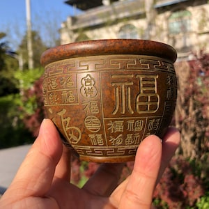 Könnte beinhalten: Eine bronzefarbene, runde dekorative Schale mit geprägten chinesischen Schriftzeichen. Die Schale hat einen breiten Rand und wird in der Hand einer Person gehalten. Die Schriftzeichen sind goldfarben und in vertikalen Spalten um die Schale angeordnet.