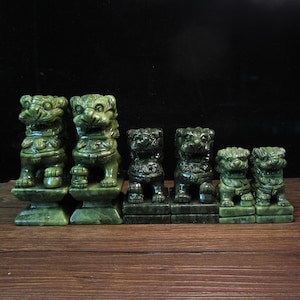 Può includere: Una collezione di sei statue di cani Foo in giada scolpita di varie dimensioni. Le statue sono di un colore verde intenso e sono esposte su una superficie di legno. I cani Foo sono in posizione seduta con tratti del viso dettagliati.