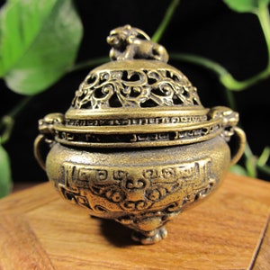 May include: A small, antique brass incense burner with intricate openwork on the lid. The lid is topped with a small lion figure. The burner has a detailed design around the base and handles on either side. The overall color is a warm, aged brass.