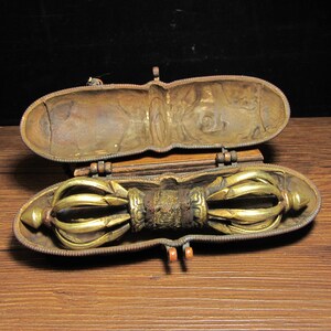 Peut inclure: Un récipient en laiton antique avec un couvercle ouvert, révélant un vajra doré et orné. Le récipient est de forme ovale avec un couvercle à charnière et une base en bois. Le vajra est un design symétrique et détaillé.