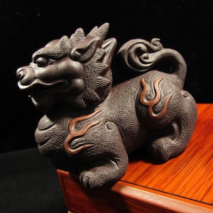 May include: A dark brown, intricately carved figurine of a mythical creature, possibly a guardian lion. The sculpture features detailed textures and stylized flame-like accents in a lighter brown color. The creature is posed in a seated position.