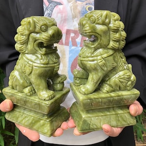 Könnte beinhalten: Zwei grüne Jade-Löwenstatuen, jeweils auf einem quadratischen Sockel sitzend. Die Löwen haben detaillierte Mähnen, offene Münder und sind mit aufwendigen Designs geschnitzt. Die Statuen sind hellgrün.