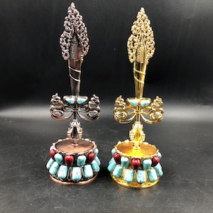 Puede incluir: Dos objetos decorativos ornamentados. Uno es de color bronce, el otro dorado. Cada uno tiene una parte superior decorativa, una base de piedras turquesas y rojas y un diseño central con detalles turquesas. Los objetos están sobre un fondo oscuro.