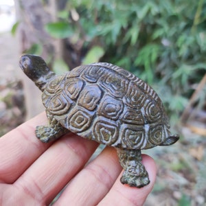 May include: A bronze-colored turtle figurine with a detailed shell pattern. The turtle's head is raised, and its legs are extended. The figurine has an aged appearance, suggesting it may be an antique or vintage piece.