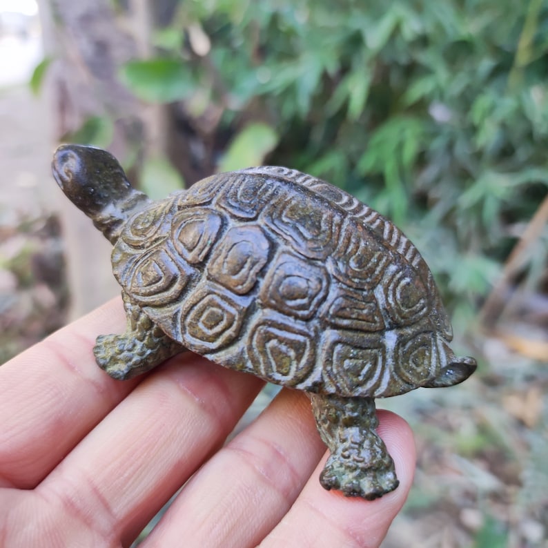 May include: A bronze-colored turtle figurine with a detailed shell pattern. The turtle's head is raised, and its legs are extended. The figurine has an aged appearance, suggesting it may be an antique or vintage piece.
