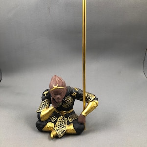 Puede incluir: Figura detallada de un mono sentado en una pose contemplativa, adornado con una armadura negra y dorada. El mono sostiene un bastón dorado. La figura tiene una cara marrón y una diadema dorada. El diseño general es intrincado y detallado.