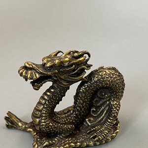May include: A detailed brass dragon figurine with a textured, scaled body and intricate facial features. The dragon is posed in a coiled position, with its head raised. The figurine has a weathered, antique finish. The dragon is a decorative item.