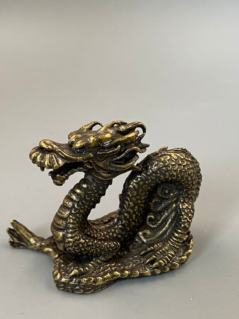 May include: A detailed brass dragon figurine with a textured, scaled body and intricate facial features. The dragon is posed in a coiled position, with its head raised. The figurine has a weathered, antique finish. The dragon is a decorative item.