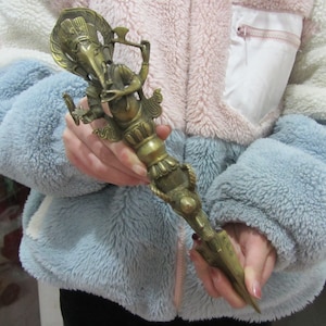 May include: An antique bronze ritual object, featuring a deity with an elephant head and multiple arms. The object has a pointed end and detailed carvings. It is held against a backdrop of a fluffy, multi-coloured jacket.