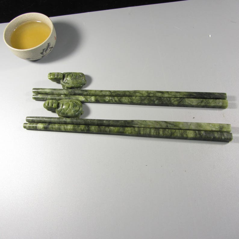Pode incluir: Dois pares de pauzinhos de m&aacute;rmore verde, cada um com um topo em forma de tartaruga. Uma pequena ch&aacute;vena de ch&aacute; esbranqui&ccedil;ada com um desenho floral e ch&aacute; no interior est&aacute; no canto superior esquerdo. Os pauzinhos t&ecirc;m um padr&atilde;o verde marmoreado.