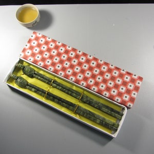 Pode incluir: Um conjunto de pauzinhos de pedra verde com pontas em forma de tartaruga, apresentado numa caixa branca com um padr&atilde;o floral vermelho e branco. Os pauzinhos s&atilde;o presos por bandas douradas. Uma pequena ch&aacute;vena de ch&aacute; no fundo.