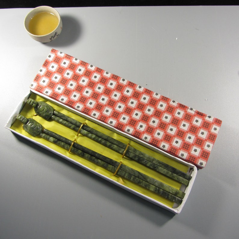 Pode incluir: Um conjunto de pauzinhos de pedra verde com pontas em forma de tartaruga, apresentado numa caixa branca com um padr&atilde;o floral vermelho e branco. Os pauzinhos s&atilde;o presos por bandas douradas. Uma pequena ch&aacute;vena de ch&aacute; no fundo.