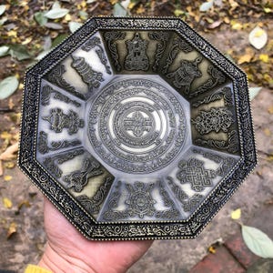 Può includere: Vassoio ottagonale in metallo in stile antico con intricati disegni in rilievo. Il vassoio presenta un emblema circolare centrale circondato da immagini e iscrizioni più piccole e dettagliate. La combinazione di colori è un mix di bronzo e argento, con un bordo decorativo.
