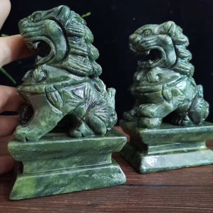 Può includere: Due statue di leoni in giada verde. I leoni sono scolpiti con criniere dettagliate, bocche aperte e seduti su basi rettangolari. Le statue sono di un colore verde intenso con variazioni di tonalità.