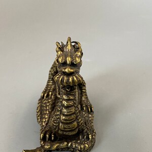 May include: A detailed brass dragon figurine with a textured surface. The dragon is depicted in a seated position, with its head raised and its mouth slightly open. The figurine has intricate details, including scales, claws, and a spiky mane. The dragon is a golden-brown colour.
