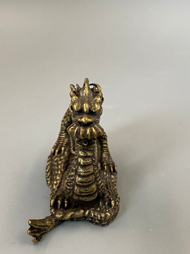 May include: A detailed brass dragon figurine with a textured surface. The dragon is depicted in a seated position, with its head raised and its mouth slightly open. The figurine has intricate details, including scales, claws, and a spiky mane. The dragon is a golden-brown colour.