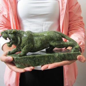 Op de afbeelding: Een gebeeldhouwde jade tijgersculptuur in een brullende pose. De groene tijger staat op een rechthoekige basis, met gedetailleerde gravures van de vacht en gezichtskenmerken. De sculptuur wordt voor een lichte achtergrond gehouden.