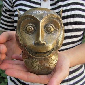 May include: A bronze-colored sculpture of a monkey's head with a smiling expression. The sculpture has detailed features, including large eyes, a prominent nose, and textured fur. The monkey's head is held in open hands.