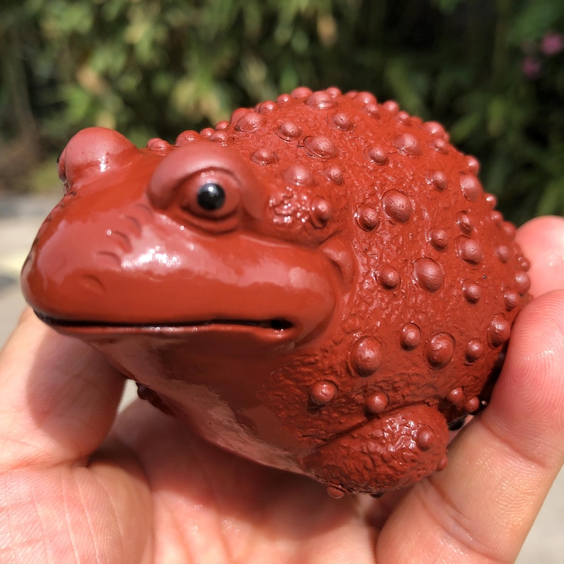 May include: A reddish-brown frog figurine with a textured surface. The frog has a detailed design with visible bumps and a smooth, glossy finish. The figurine is held in a hand, showcasing its size and shape. The frog's eyes are black.