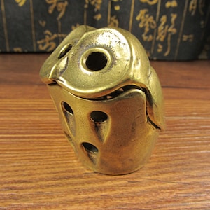 May include: A brass owl ornament with a hollow body and large eye sockets. The owl has a textured surface and is on a wooden surface. The figurine is a decorative item.