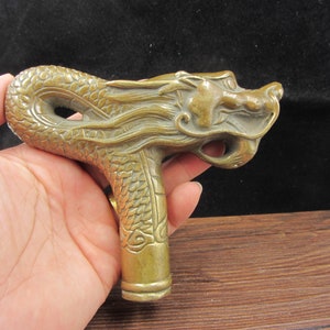 May include: A brass-colored dragon-head cane handle. The handle is intricately detailed with scales and a dragon's face. The handle is held in a hand, with a dark background.