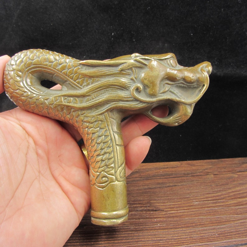 May include: A brass-colored dragon-head cane handle. The handle is intricately detailed with scales and a dragon's face. The handle is held in a hand, with a dark background.