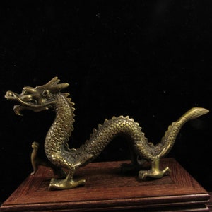 May include: A detailed brass dragon figurine with a curved body, standing on four legs, and a detailed head. The dragon is positioned on a small, rectangular wooden base. The background is dark.