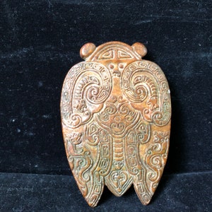 May include: A brown, intricately carved jade pendant with a unique shape. The pendant features detailed patterns and designs, including swirling lines and geometric shapes. The overall design is symmetrical and has a three-pronged bottom.