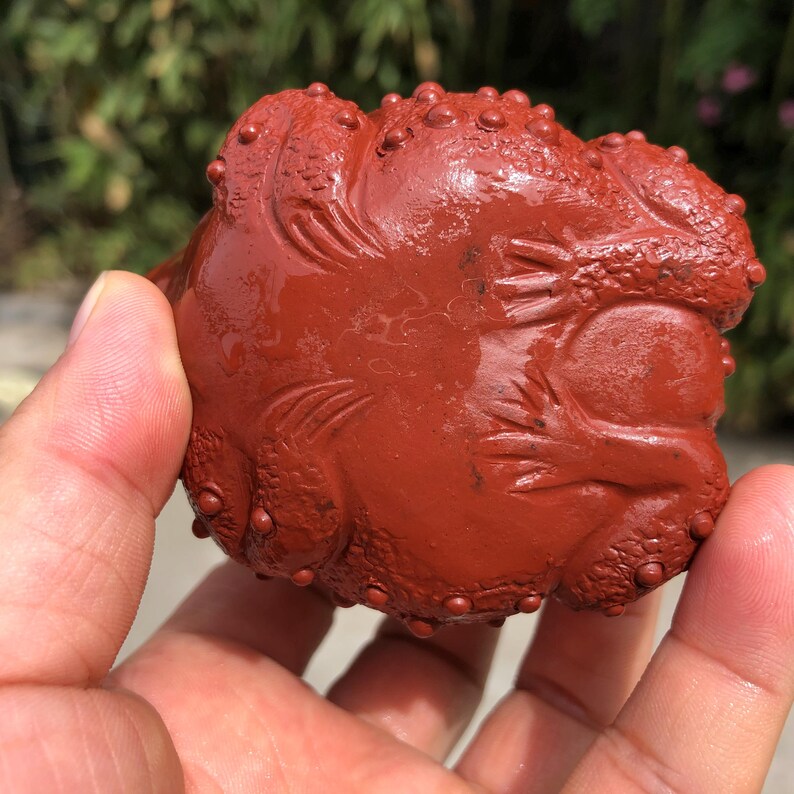 May include: A reddish-brown, carved stone sculpture of a toad. The toad has a textured surface with small bumps and detailed carvings. The sculpture is held in a hand, with a blurred green background.