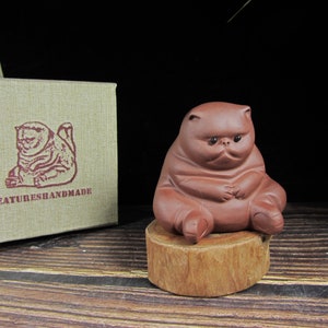 Puede incluir: Figura de gato de cerámica marrón sentada sobre una pequeña base de madera. El gato tiene una expresión de mal humor y está sentado con las piernas extendidas. Una caja con el texto "CREATURESHANDMADE" está en el fondo.