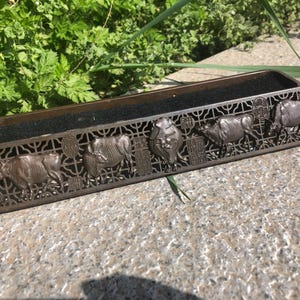 May include: A rectangular, bronze-colored incense burner with a decorative lattice design. The front features detailed relief carvings of oxen and a central emblem. The interior is lined with a dark, possibly felt, material.