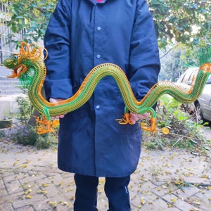 May include: A handcrafted wire dragon sculpture in shades of green and orange. The dragon is held by a person, showcasing its detailed design and flowing form. The sculpture features intricate wire weaving, creating a unique and artistic representation of the mythical creature.
