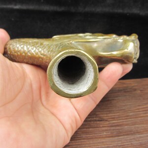 May include: A brass dragon-shaped pipe with a detailed, textured body and head. The pipe's bowl is a circular opening with a white interior. The dragon's head is at the end of the pipe, and the overall design is antique.