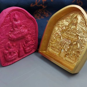 Puede incluir: Un molde rectangular rosa con un relieve detallado de una figura sentada y otras figuras. Junto a él, un molde dorado con un diseño de relieve similar. Los moldes son probablemente para crear artículos decorativos.