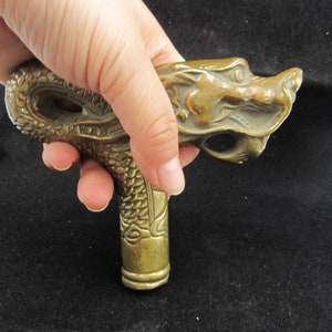 May include: A bronze-colored cane handle with a dragon design. The handle features a detailed dragon head and scales, with a cylindrical base. The dragon's head is facing right, and the handle is held in a hand.