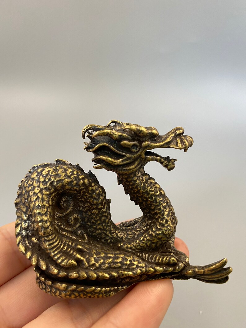 May include: A detailed brass dragon figurine with a textured body and intricate facial features. The dragon is posed in a curved position, with its head raised and mouth open. The figurine has a vintage appearance.