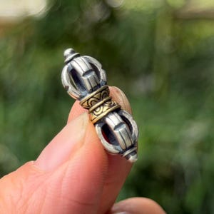 Pode incluir: Um pequeno pingente vajra metálico com um corpo de tom prateado e uma faixa central de tom dourado. O objeto tem um design simétrico com extremidades pontiagudas e padrões gravados intrincados na seção central, com um acabamento polido.