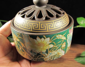 May include: A ceramic incense burner with a dark green base and a decorative lid. The base features a floral design with gold accents. The lid has a geometric pattern and a small knob on top.