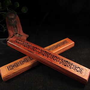 May include: Two wooden incense holders with detailed cut-out patterns. One is a deep reddish-brown, the other a lighter brown. The rectangular holders display decorative designs. The dark background accentuates the natural colours of the wood.