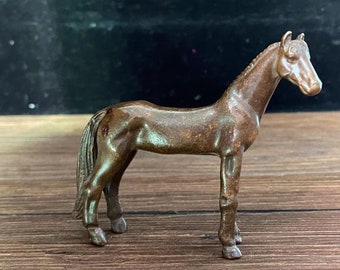 以下が含まれることがあります： ブロンズ色の馬の置物。馬は頭をわずかに右に向けて立っています。この置物には、なびくたてがみや尾など、詳細な特徴があります。馬は木製の表面に置かれています。