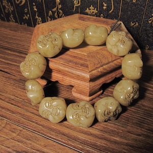 May include: A bracelet composed of ten light beige, translucent stone beads, each intricately carved with a stylized, smiling face featuring prominent eyebrows and a wide mouth, strung together on a dark cord in a circular arrangement.
