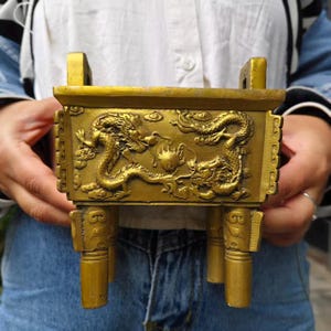 Puede incluir: Un quemador de incienso rectangular de latón de estilo antiguo. Los lados presentan detalladas tallas en relieve de dragones. El quemador tiene cuatro patas cilíndricas y dos asas. El color general es un oro cálido y envejecido.